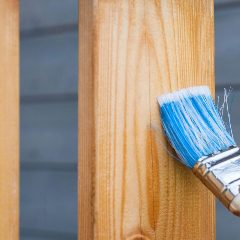 A hand painting a wooden fence plank with a blue paintbrush outdoors. Perfect for DIY and renovation themes.